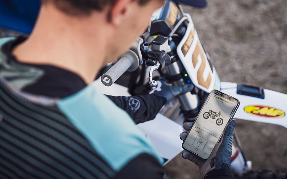 Piloto de motocross sosteniendo un teléfono con la Husqvarna Motorcycles App junto a su moto.
